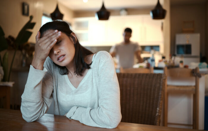 A woman sits at a table in a kitchen, holding her forehead with a pained expression, appearing stressed or overwhelmed. In the background, a man stands near the kitchen counter, slightly blurred, suggesting a domestic setting and a tense or difficult situation.